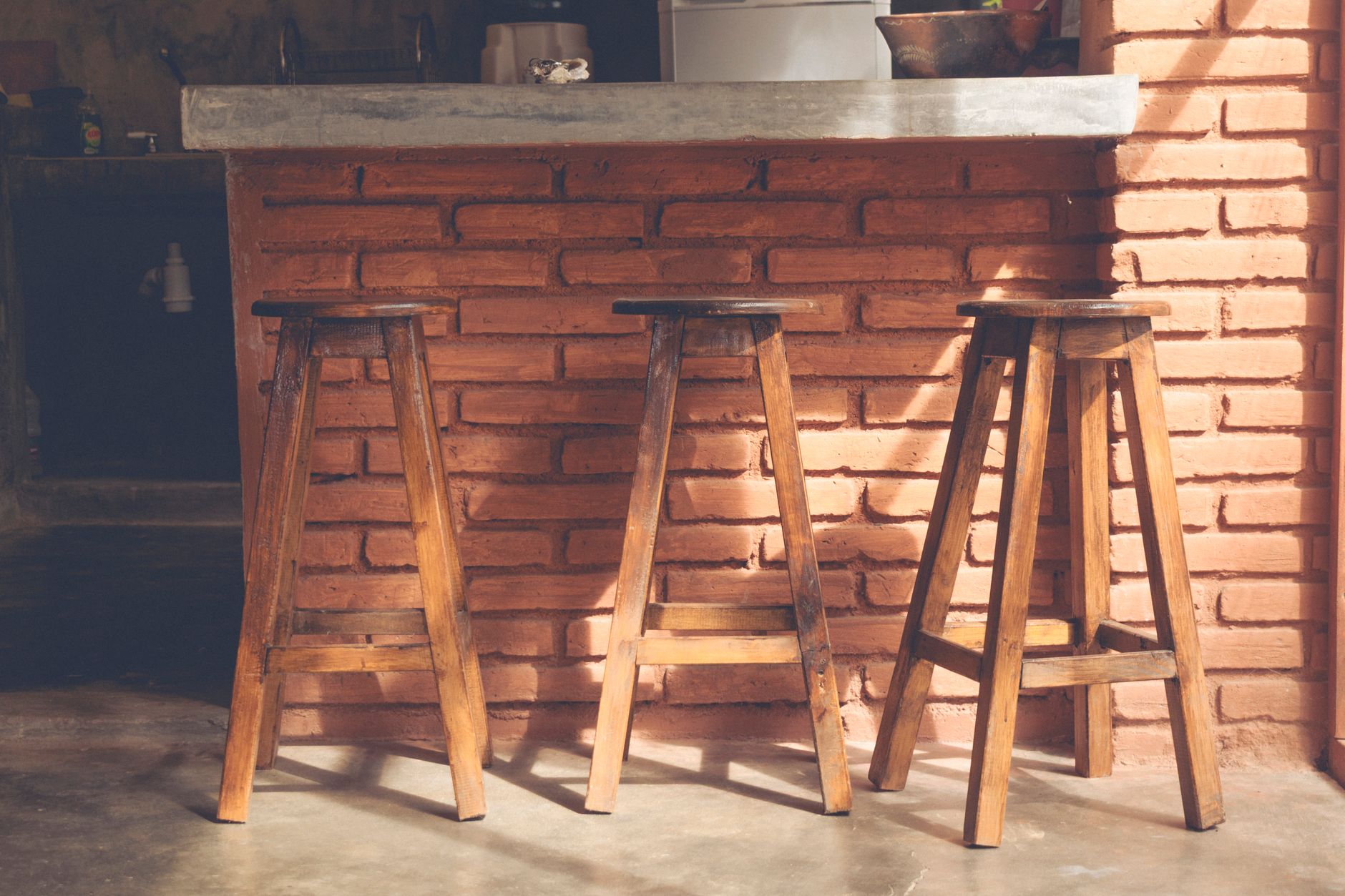 Three wooden bar stools in a rustic kitchen setting with brick walls and natural lighting.