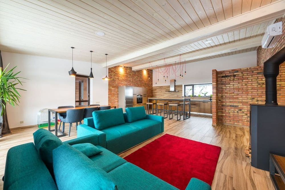 Spacious modern living room featuring brick walls, teal sofa, and red rug.
