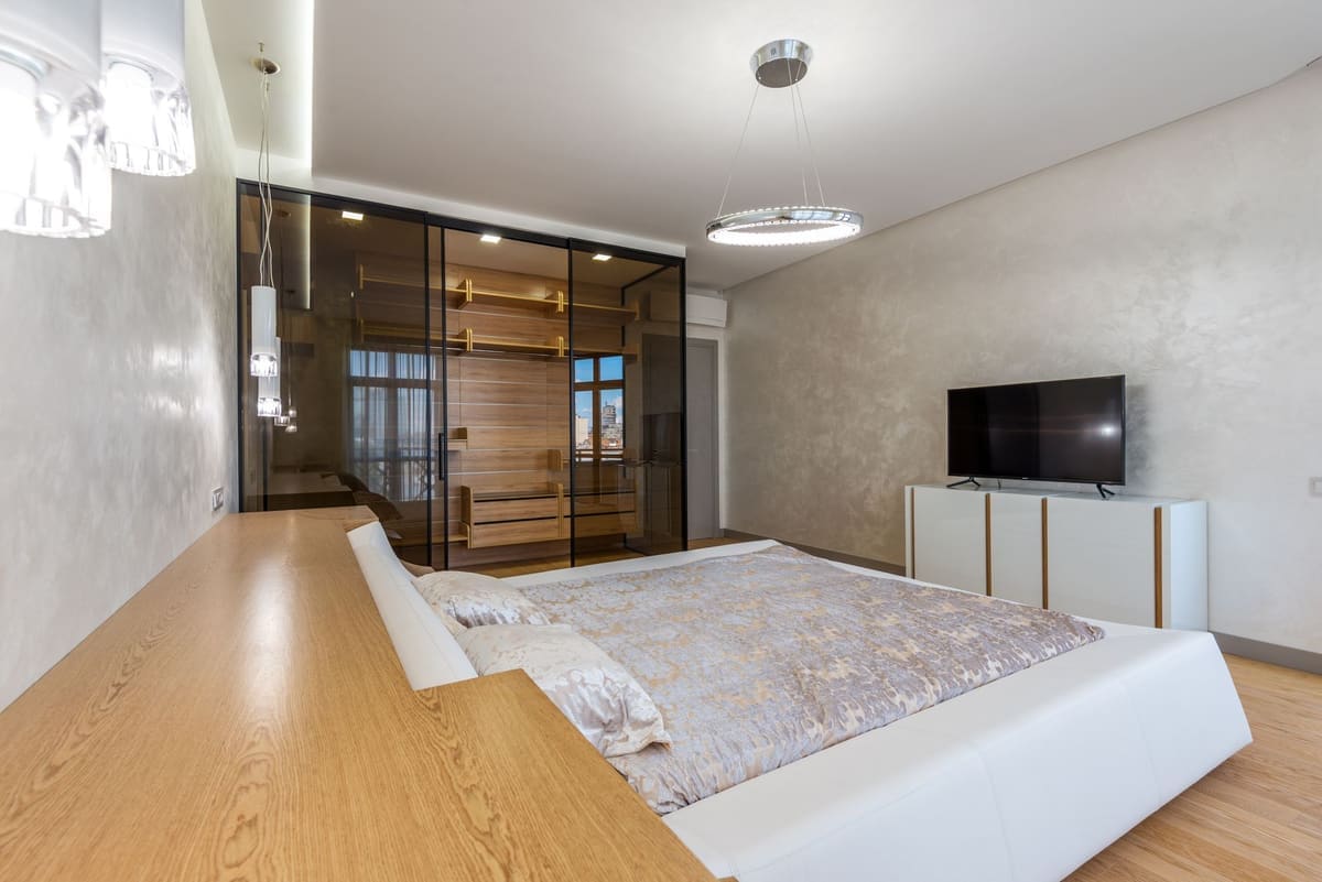 Bed with pillows placed against wall with TV on cupboard in spacious bedroom and glowing chandelier with wardrobe in apartment