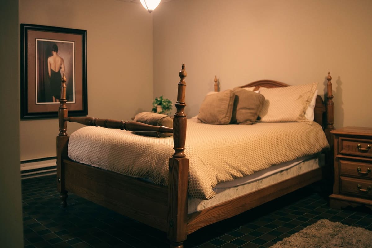 Warmly lit bedroom with wooden furniture, a made bed, and a classic painting on the wall.