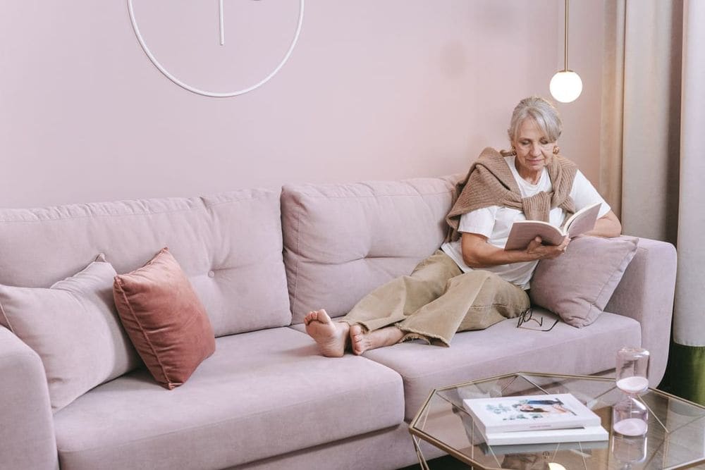 Senior woman relaxing and reading a book at home on a comfortable pink sofa.