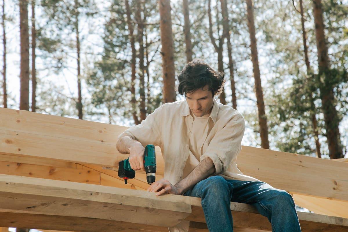 Focused carpenter uses power drill on wooden beams in an outdoor setting, surrounded by trees.