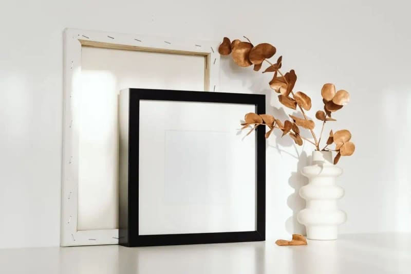 A minimalist home setting with empty frames and a vase with dried eucalyptus branches on a white surface.