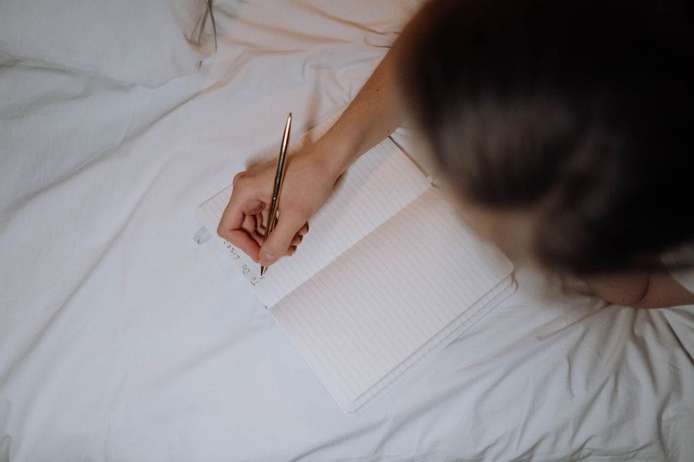 A person writing a to-do list in a notebook while sitting on a bed with white linen.