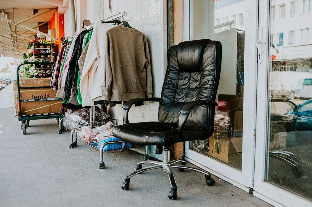 Worn black office chair outside a secondhand store with clothes and boxes nearby.