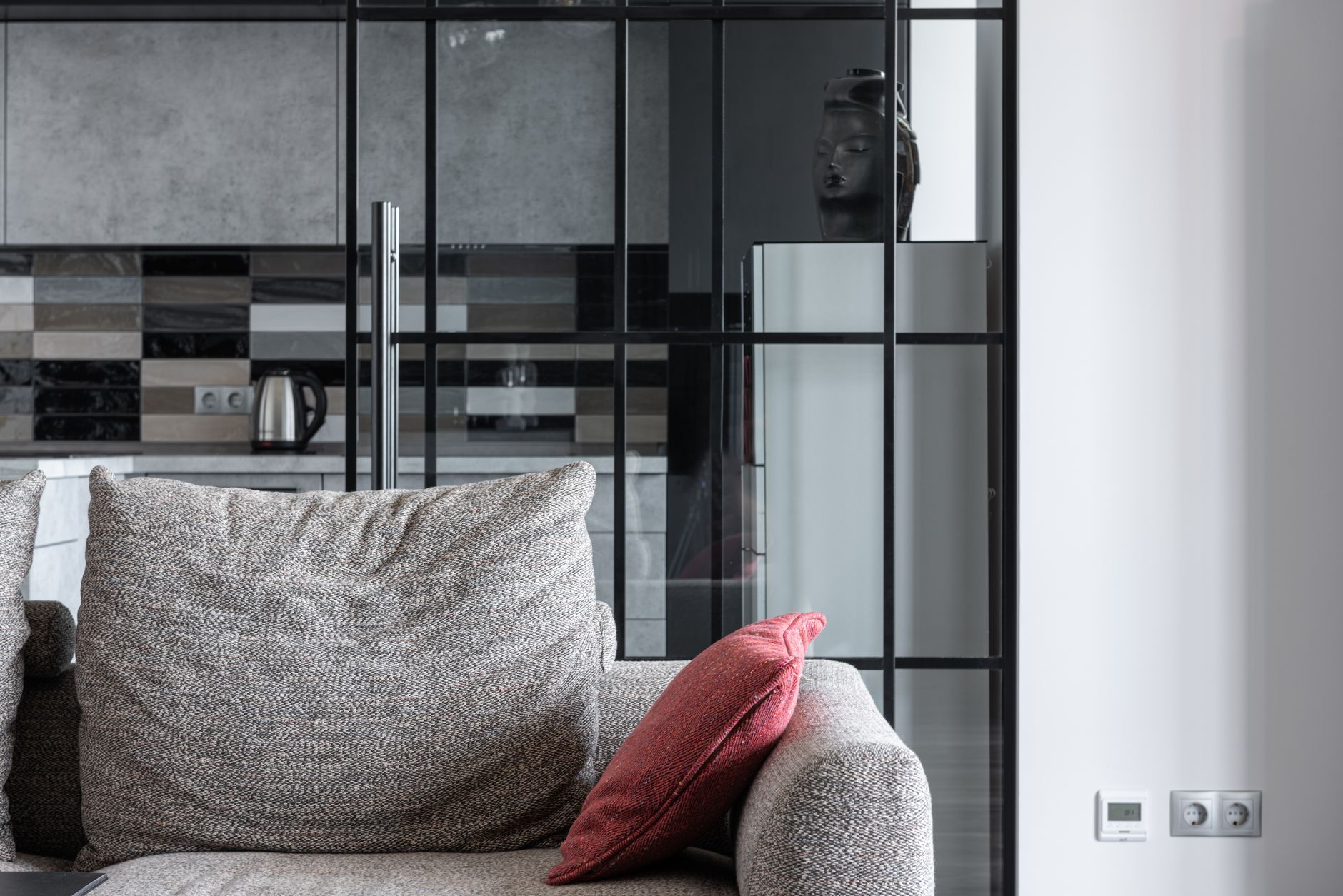 Cozy living room featuring a textured sofa with a red pillow and a modern kitchen backdrop.