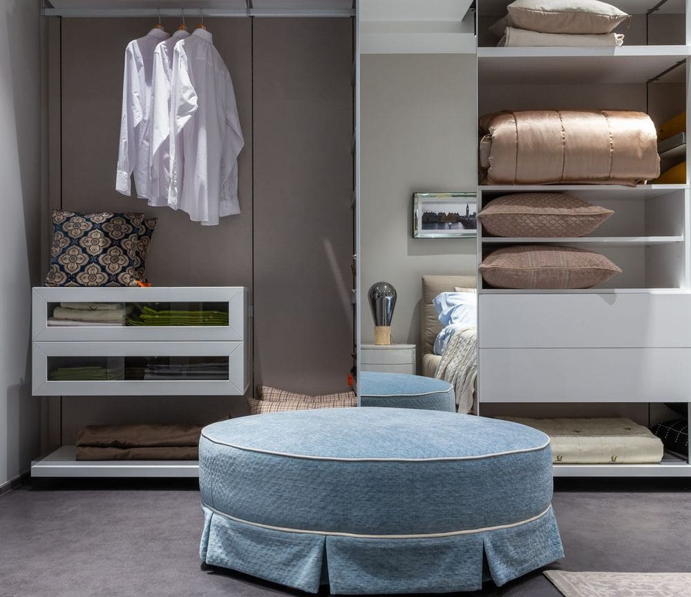 Comfortable round pouf near opened wardrobe with hanging clothes and bedding on shelves placed against mirror with reflection if bed in bedroom