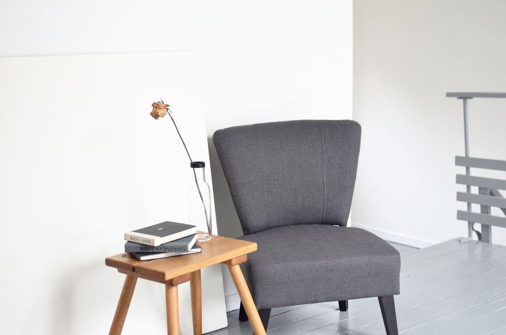 Vase with dry rose and stack of books arranged on small wooden table near comfortable armchair in modern light office