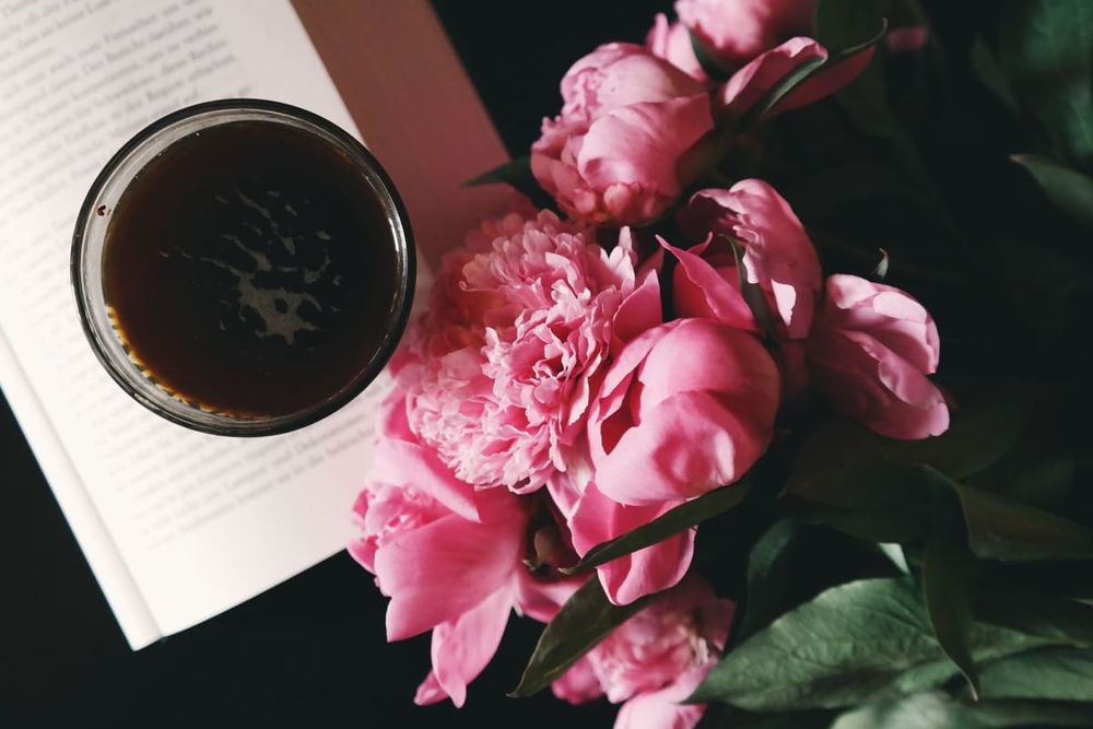 Aesthetic arrangement of pink peonies, a coffee glass and an open book, creating a cozy and artistic vibe.