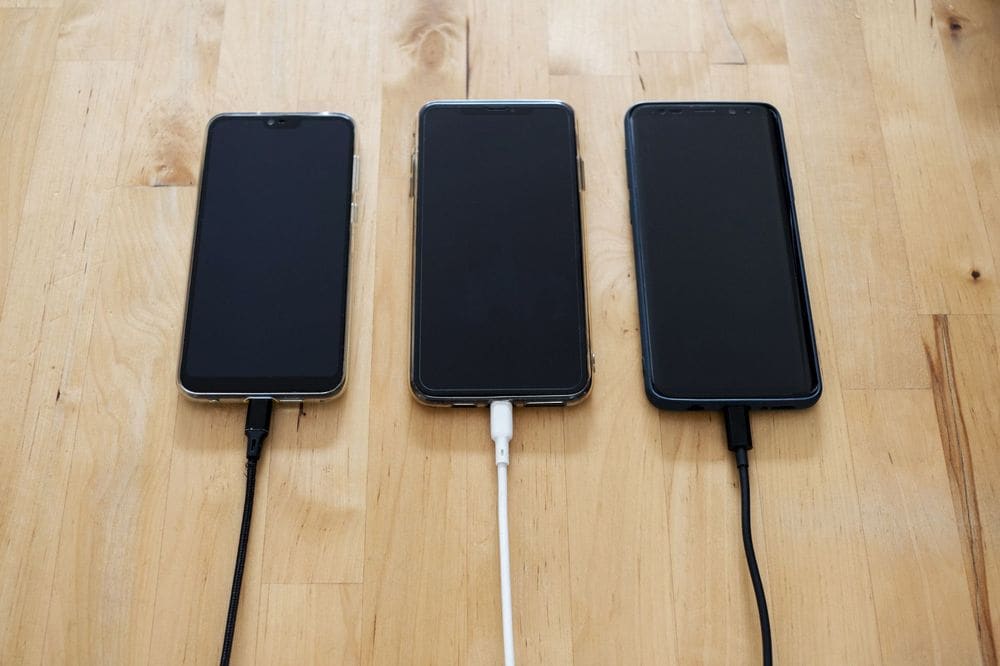 Three smartphones connected to chargers on a wooden surface, showcasing modern technology.