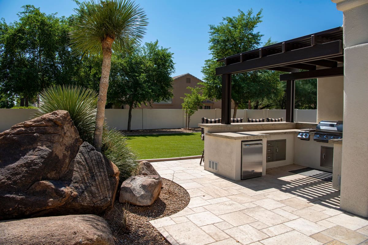 Stylish outdoor kitchen on a sunny patio with lush landscaping and clear blue sky.