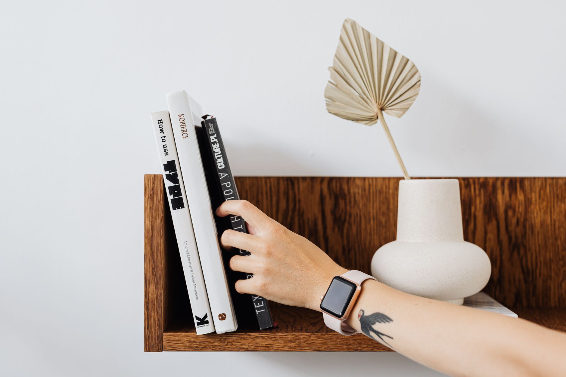 A hand reaches for books on a minimalist wooden shelf, featuring a vase with a palm leaf.